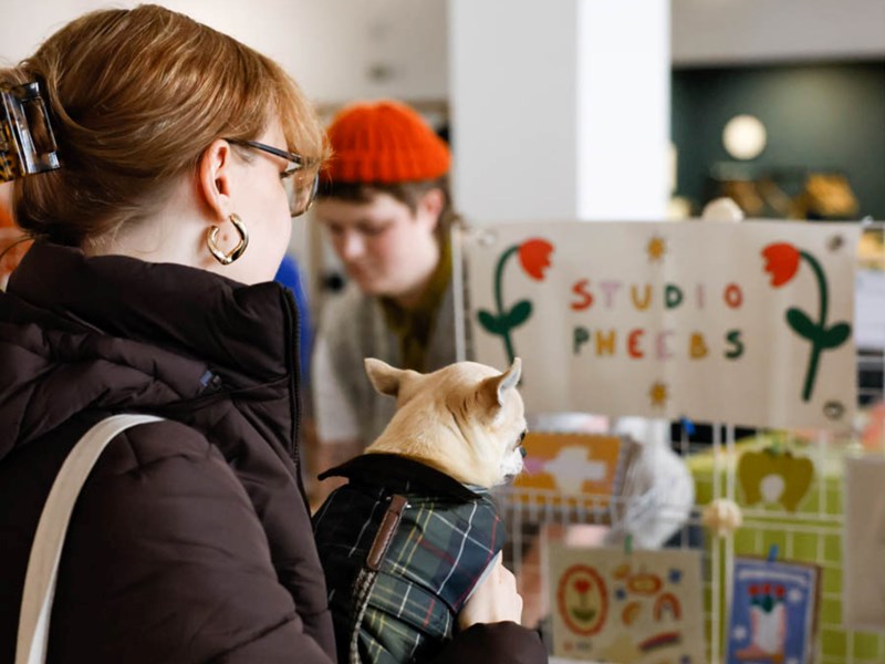 A person wearing glasses and a dark jacket holds a small dog in a plaid coat in front of a booth labeled "STUDIO PHEEBS," decorated with colorful flowers. Another person in an orange beanie stands behind the booth, which displays various art or craft items, suggesting a lively market or fair setting.