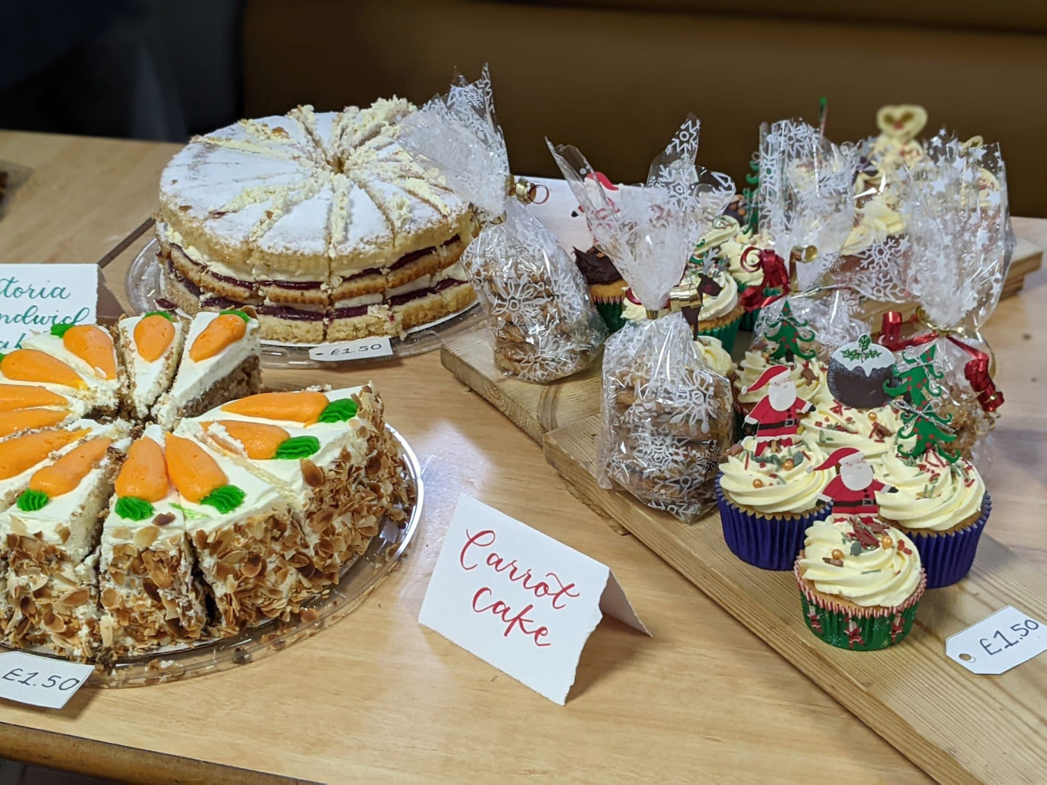 Table displaying a variety of homemade baked goods. On the left, a carrot cake topped with orange icing carrots and chopped nuts is sliced and labeled “Carrot Cake” with a price tag of £1.50 per slice. Behind it, a Victoria sponge cake with powdered sugar and jam filling is priced at £2.50 per slice. To the right, a wooden tray holds festive cupcakes decorated with Christmas-themed toppers like Santa and trees, along with clear bags of cookies tied with lace ribbon.
