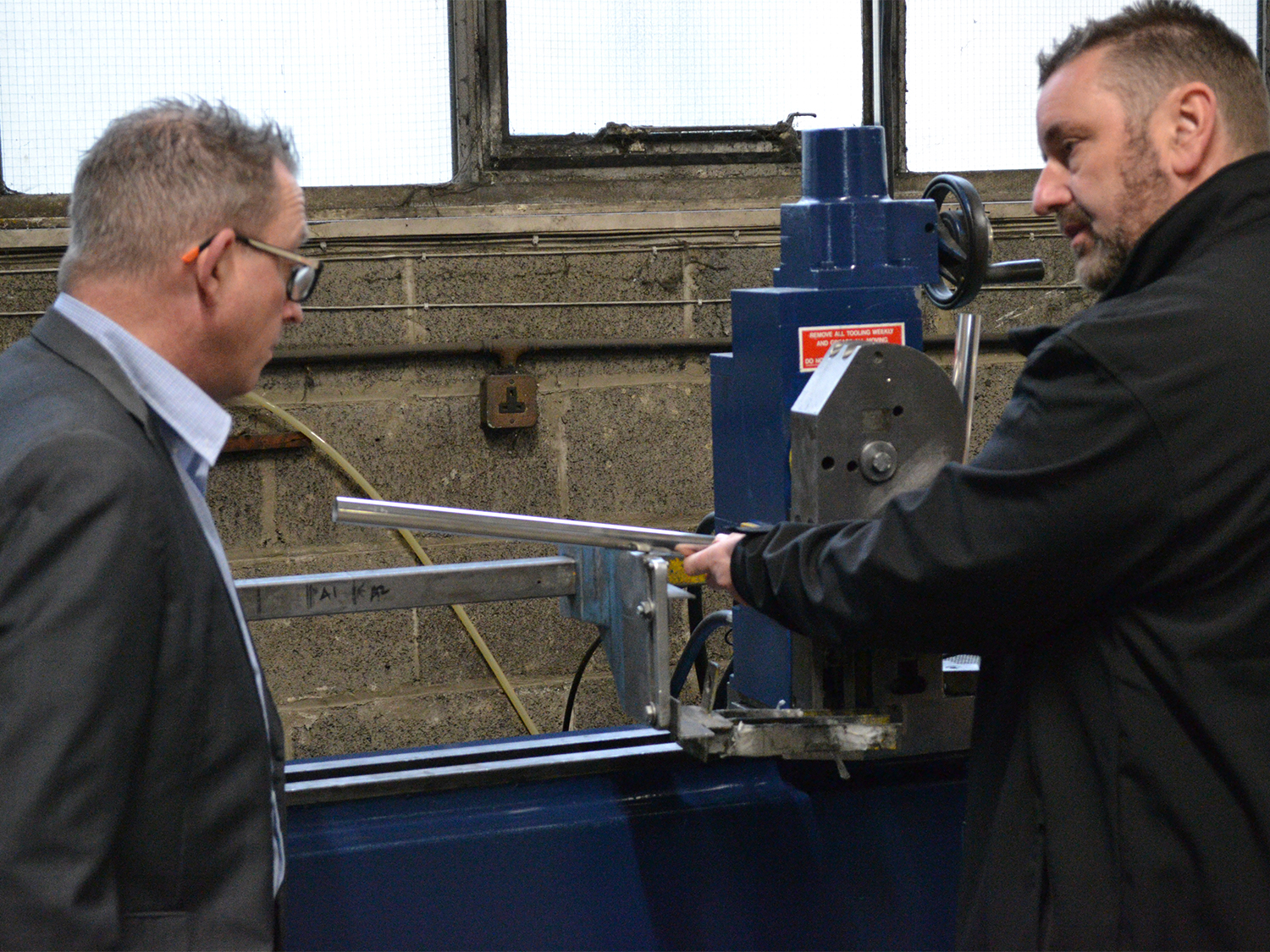 Business Sheffield's Andy Sorsby visits Blake UK. Two people standing next to a metalworking machine inside an industrial workshop. One person is holding a long metal rod positioned in the machine, while the other observes. The background shows a concrete wall, windows, and electrical wiring.