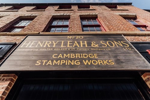 Close-up view of a large wooden sign mounted on a red-brick building. The sign reads: ‘No 20 Henry Leah & Sons Ltd, incorporating Walter Walker & Co Piercers Stampers Ltd, Cambridge Stamping Works.’ The building has tall windows and a clear blue sky above.