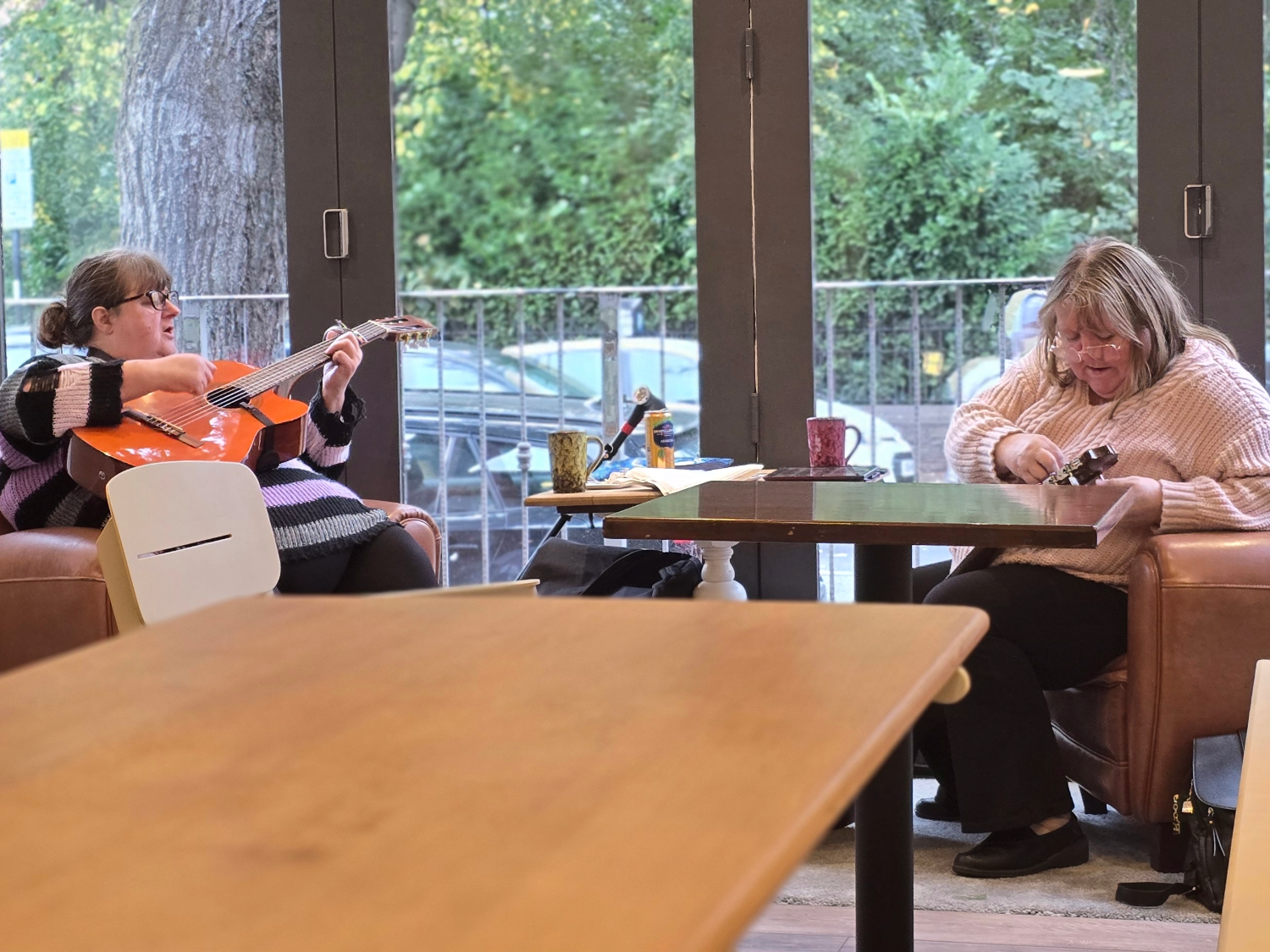 Two people seated at a table in a bright café near large glass doors with greenery visible outside. One person is holding an orange acoustic guitar, while the other is adjusting or playing a smaller string instrument. The table has mugs and papers on it, and there are chairs and wooden tables in the foreground.