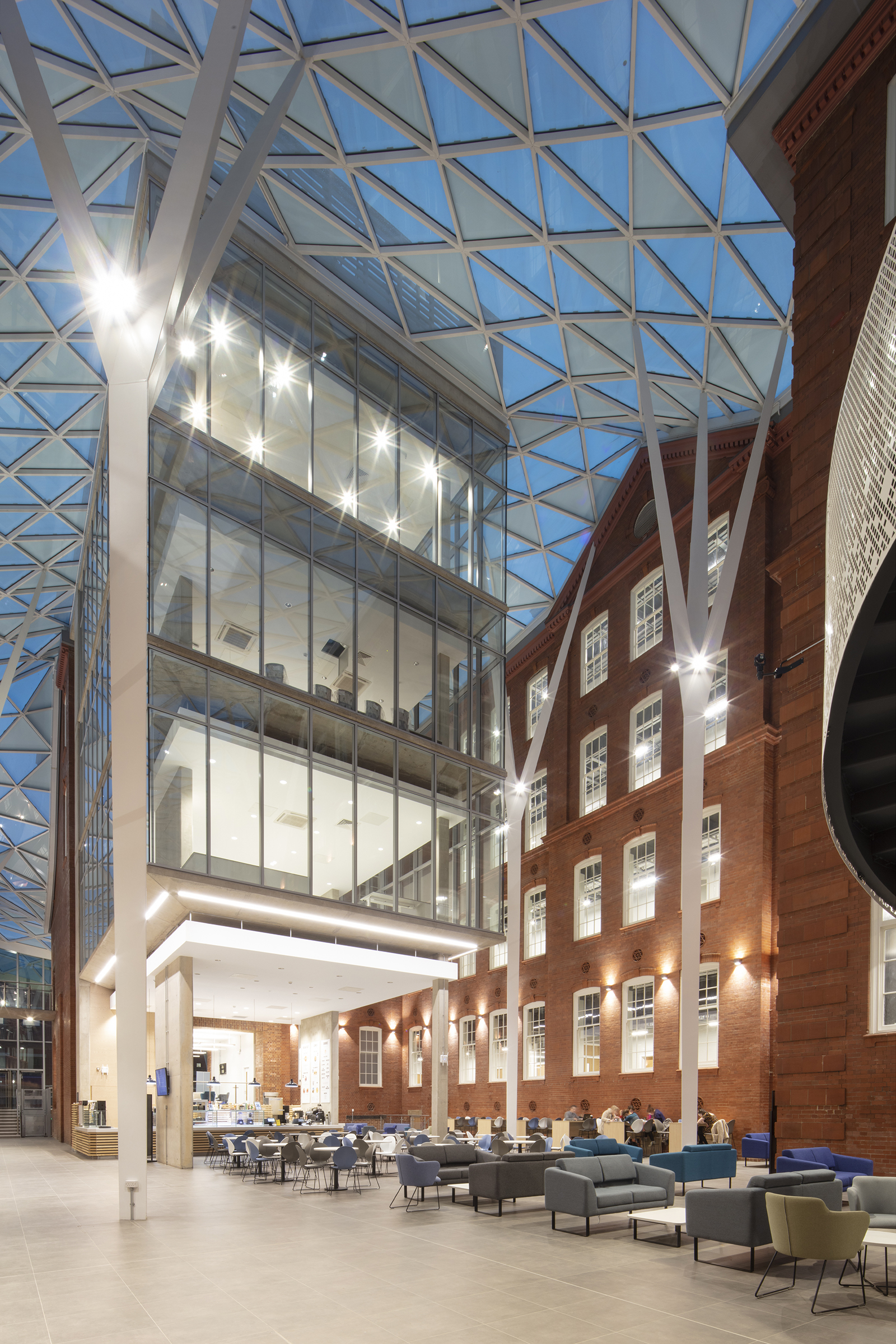 Heartspace - Modern atrium with a glass roof and steel beams connecting to a historic red-brick building. The space features multiple floors with large glass windows, bright lighting, and a seating area with tables and chairs on the ground level