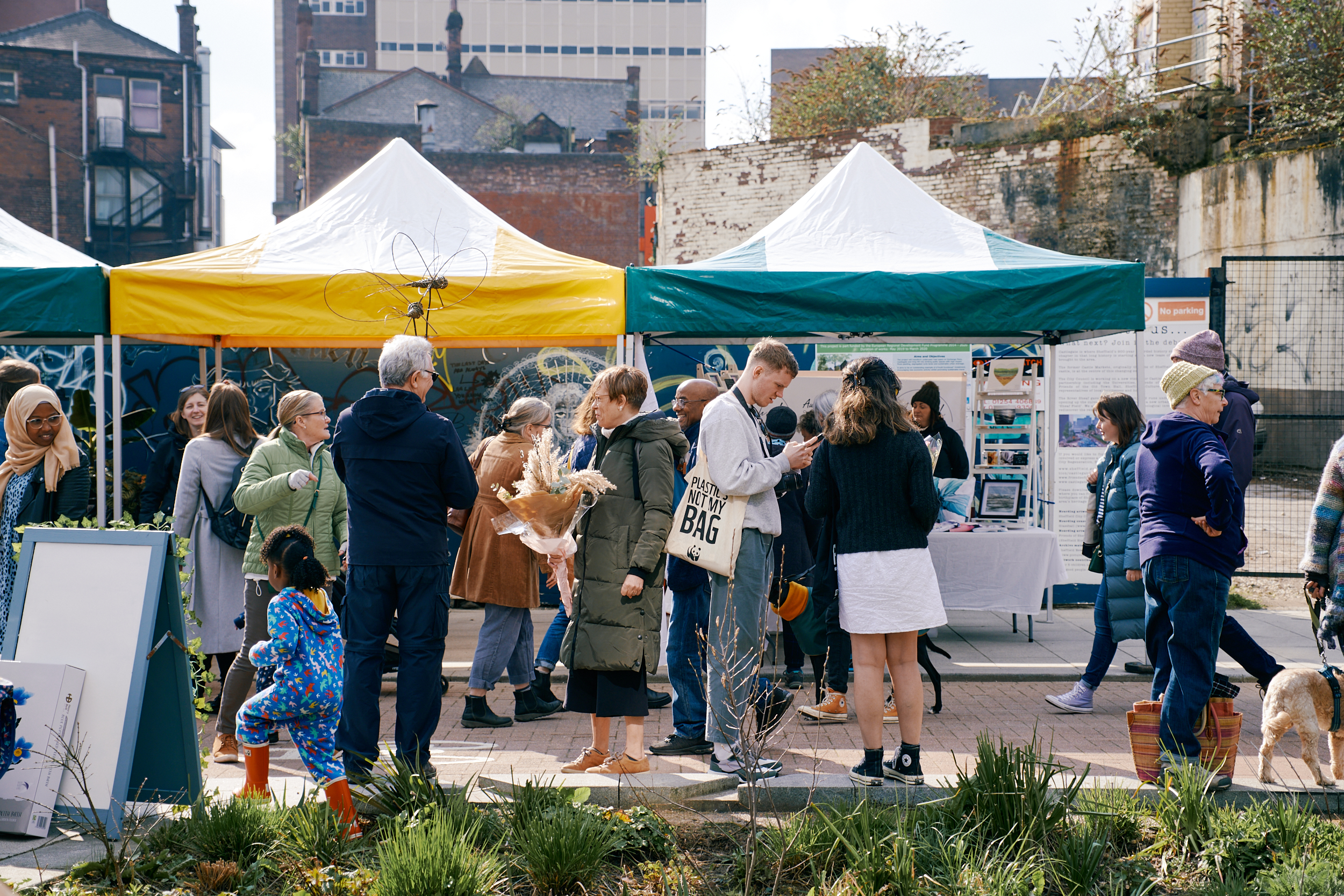 A lively outdoor market scene with people gathered around stalls covered by yellow and green canopies. Various individuals are browsing items, chatting, and holding bags or flowers. Display boards and tables with informational materials are set up under the tents. The background shows older brick buildings and a tall modern structure, while the foreground features plants and a paved area.