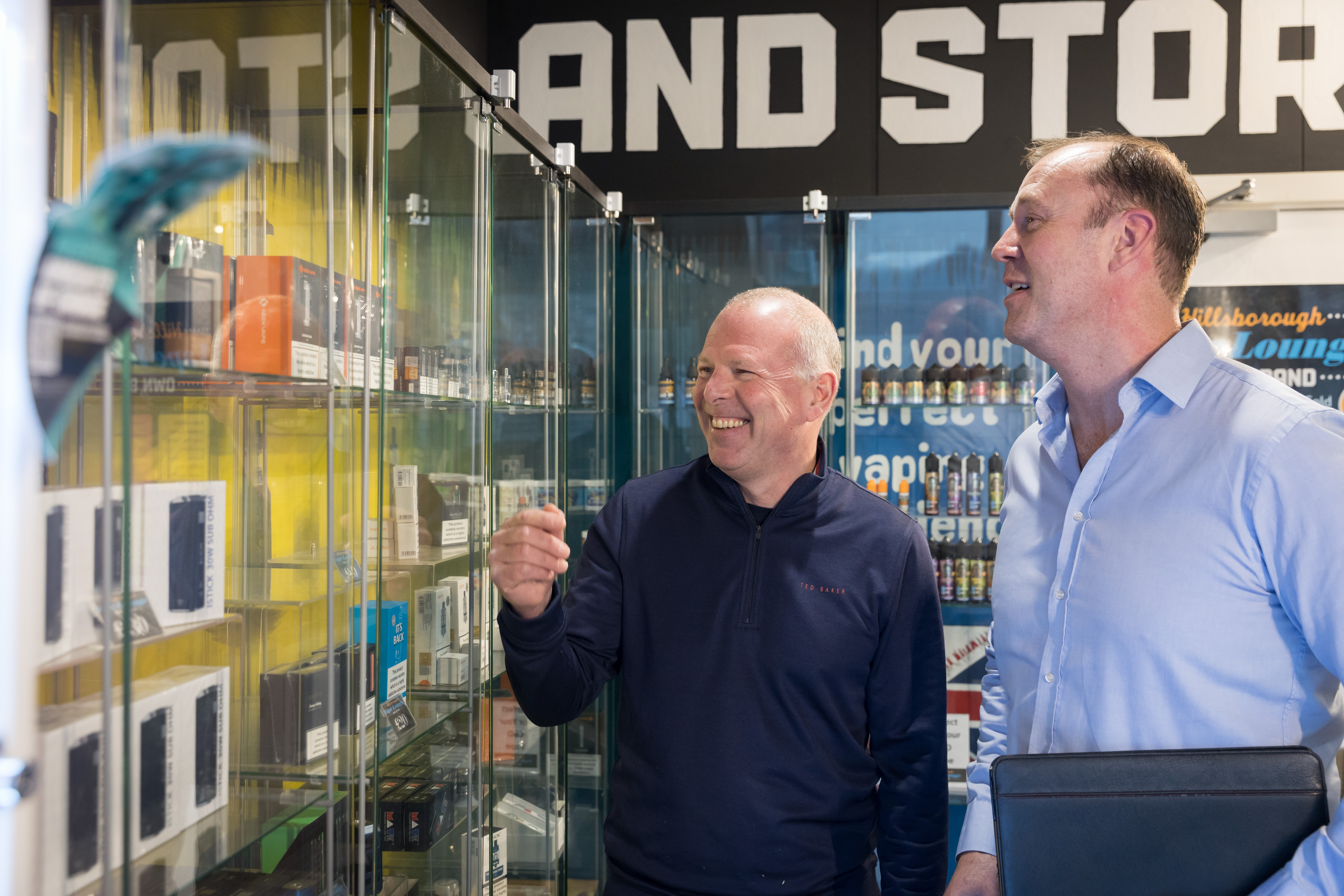 Two men, in business attire, chat in a shop while looking at glass display cabinets.