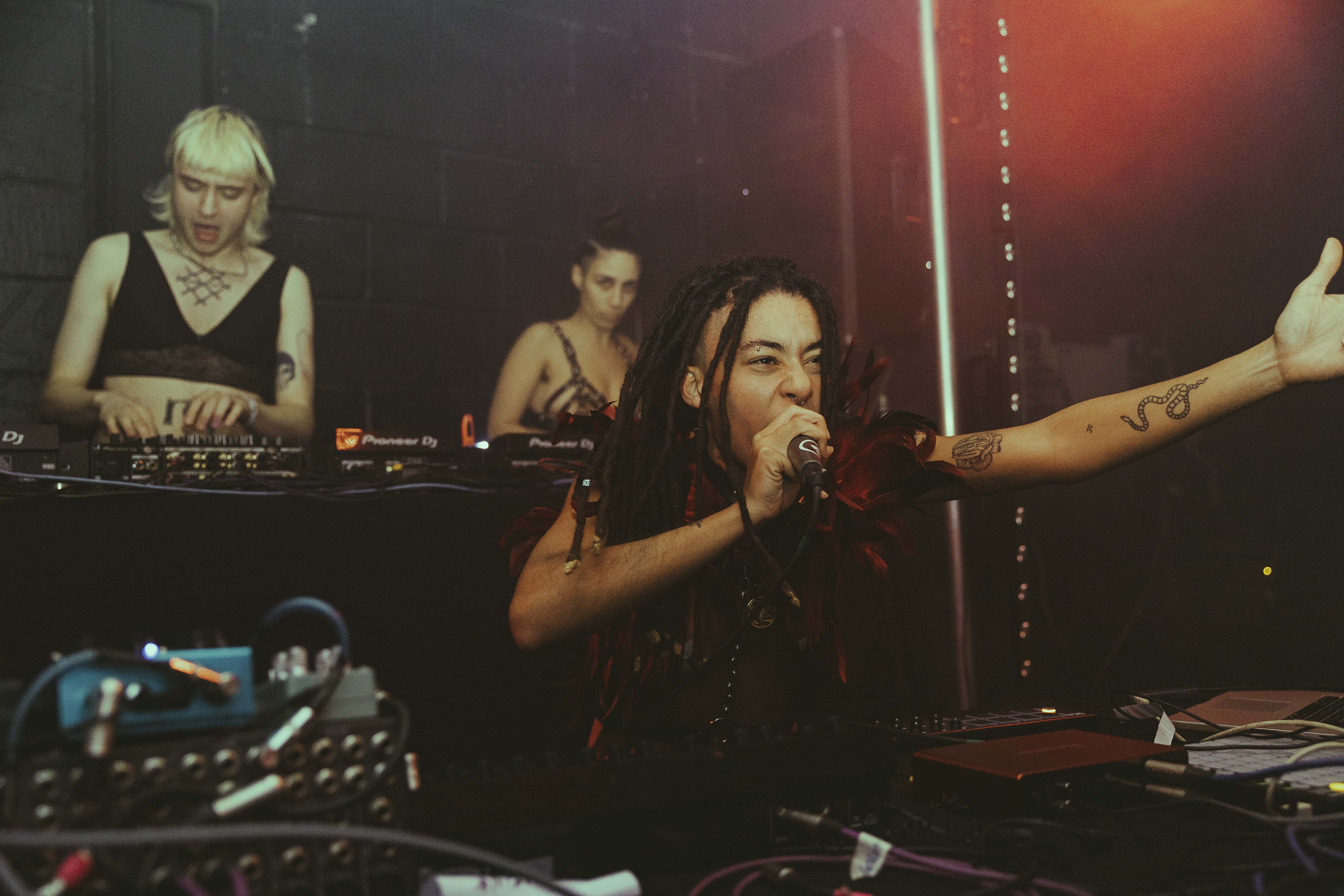 Three performers on stage at No Bounds festival. One is singing into a microphone while the other two are playing records on turntables and using a mixer.
