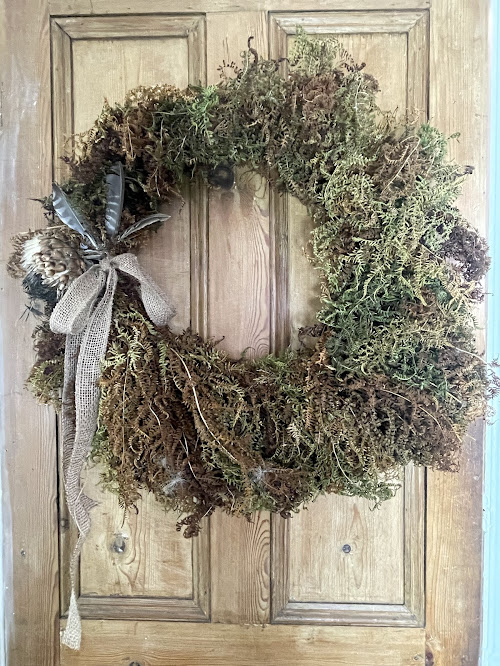 An earthy wreath composed of dried moss, ferns, and natural textures in shades of brown and green. It is decorated with a burlap ribbon and a cluster of feathers and pinecones on one side, giving it a woodland-inspired look. The wreath hangs on a wooden door.