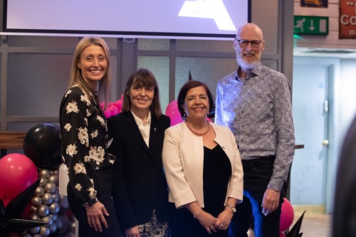Four people standing together indoors at an event, positioned in front of a backdrop with a large screen displaying a partial logo. The group is surrounded by decorative balloon arrangements in black, pink, and silver. The setting includes grey paneled walls and visible signage in the background, suggesting a formal or celebratory occasion.