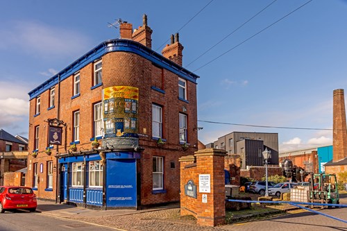 The Fat Cat pub in Kelham Island - Historic red-brick pub building with bright blue trim on a street corner, featuring a large painted sign reading ‘The Victoria Junction Inn.’ A red car is parked nearby, and industrial-style buildings and a tall chimney are visible in the background under a clear blue sky.