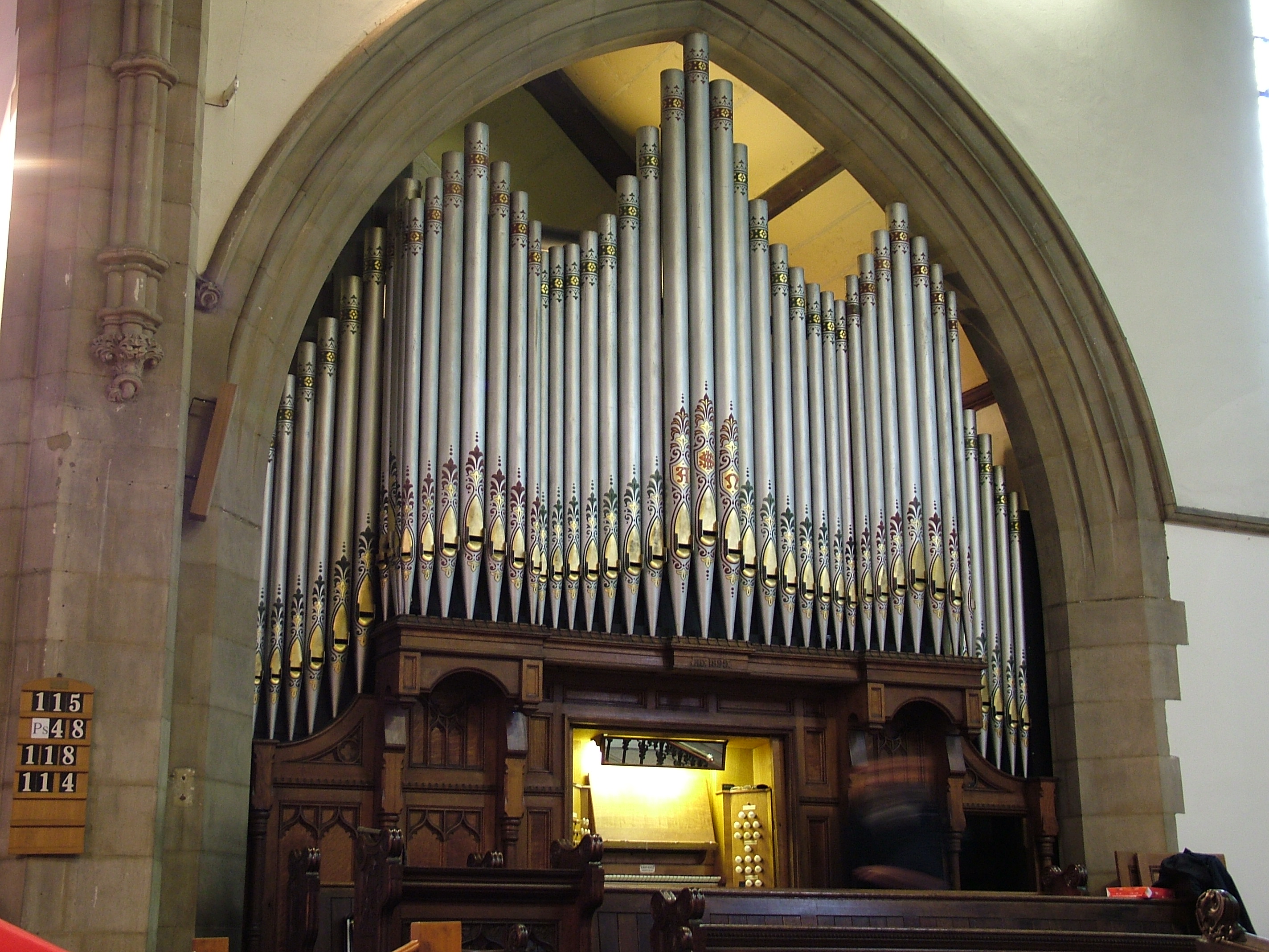 Large pipe organ inside a church, framed by a stone archway. The organ features multiple tall, silver pipes arranged in a symmetrical pattern with decorative designs near the tops. Below the pipes is a dark wooden console with ornate carvings and a keyboard area illuminated by a light. Hymn boards with numbers are mounted on the stone pillar to the left. The background shows high, light-colored walls and a vaulted ceiling.