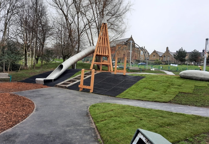 Playground with a large wooden-framed structure and a long, curved metal slide. The area is surrounded by grass, pathways, and leafless trees. Stone buildings and fencing are visible in the background under an overcast sky.