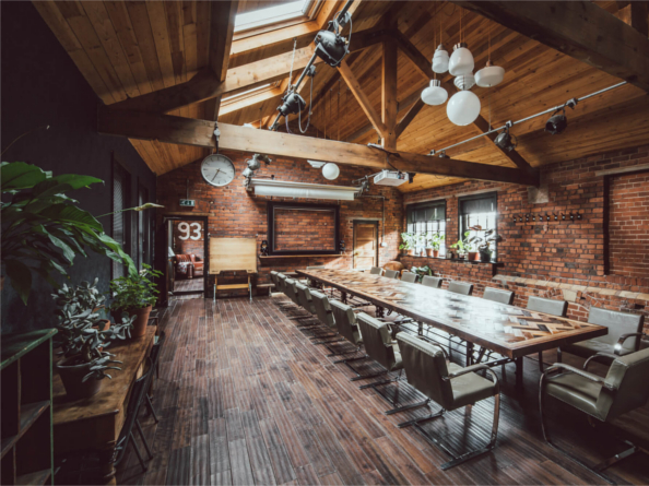 Inside The Chimney House . Spacious meeting room with exposed brick walls, wooden ceiling beams, and large skylights allowing natural light. A long rectangular table with modern chairs runs down the center of the room. Pendant lights and spotlights hang from the ceiling, and plants decorate the side tables and windowsills. A large wall clock and framed board are mounted on the far wall, adding to the rustic-industrial design.