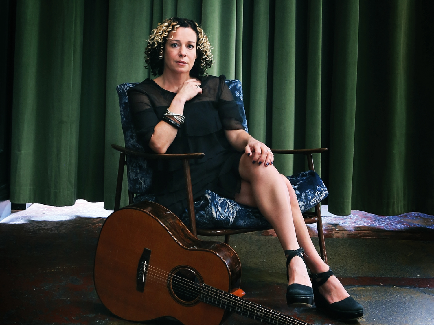 A person seated in a patterned armchair with legs crossed, wearing a black dress and black shoes. The setting includes a dark green curtain backdrop and a polished floor with a mix of colours. A wooden acoustic guitar rests on the floor in front of the chair, leaning slightly toward the person.
