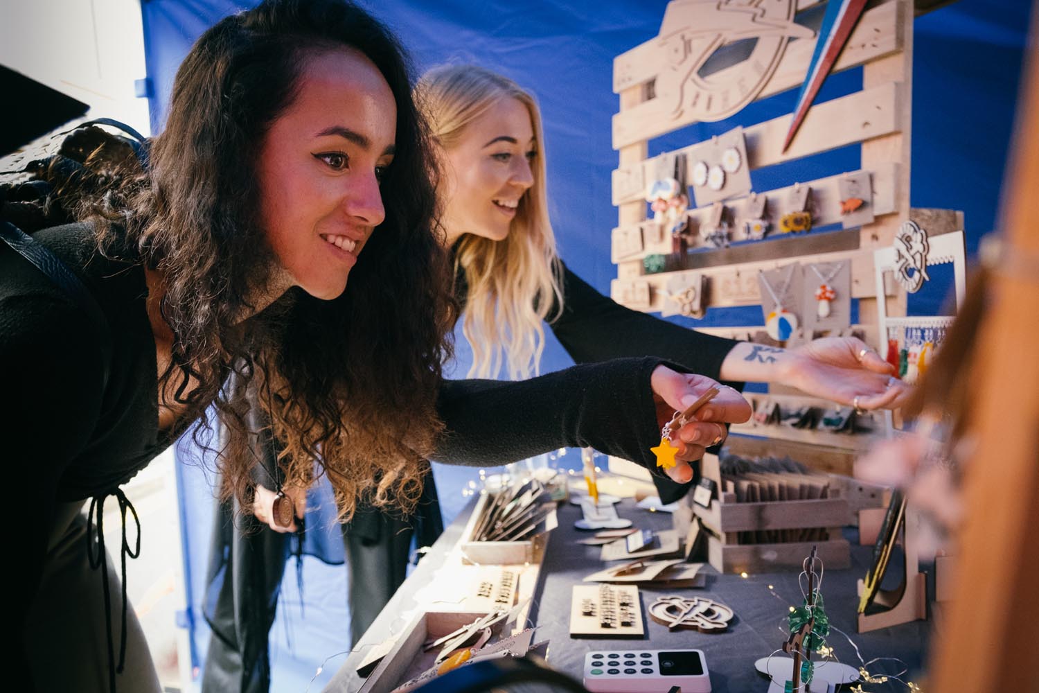 Two people browsing a craft stall displaying handmade wooden items and jewelry. The table is filled with small decorative pieces, and a wooden display rack holds various ornaments and accessories. The setting appears to be under a tent with bright lighting.