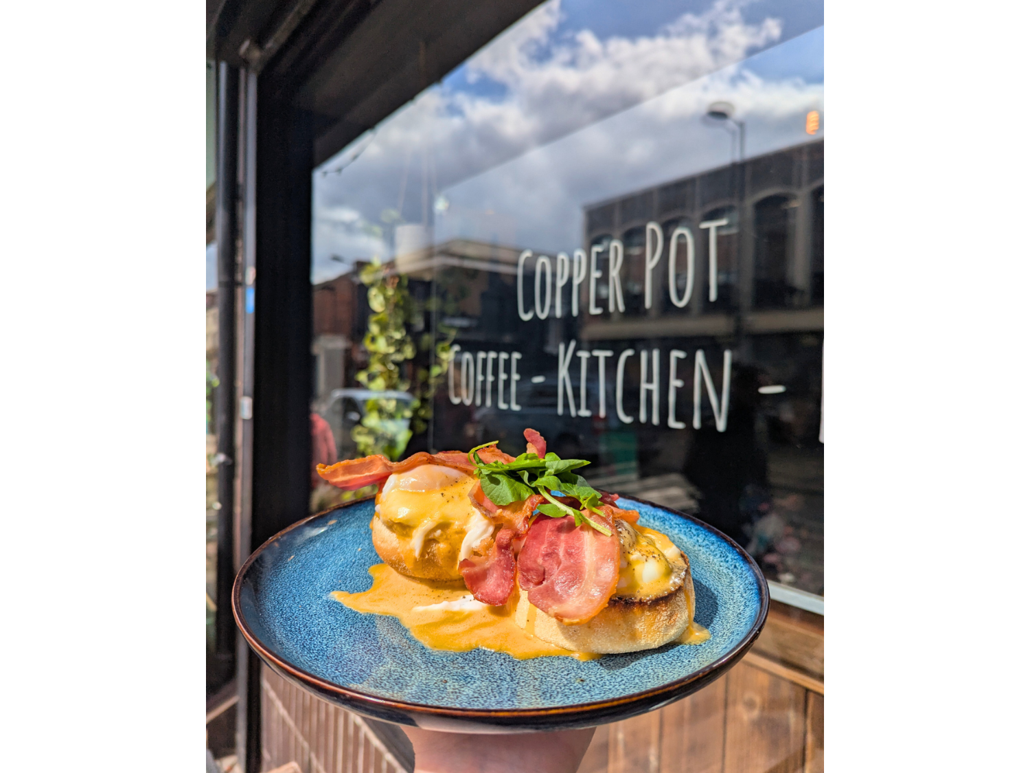Eggs Benedict with bacon, on a blue plate, being held up outside the Copper Pot Café. 