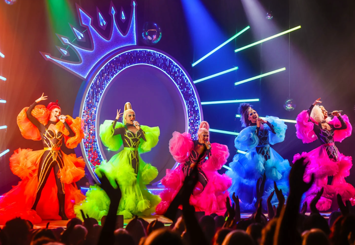 Queenz performing on stage in amazing colourful frocks against a backdrop of a huge neon letter Q with a crown perched on top.