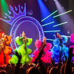 Queenz performing on stage in amazing colourful frocks against a backdrop of a huge neon letter Q with a crown perched on top.