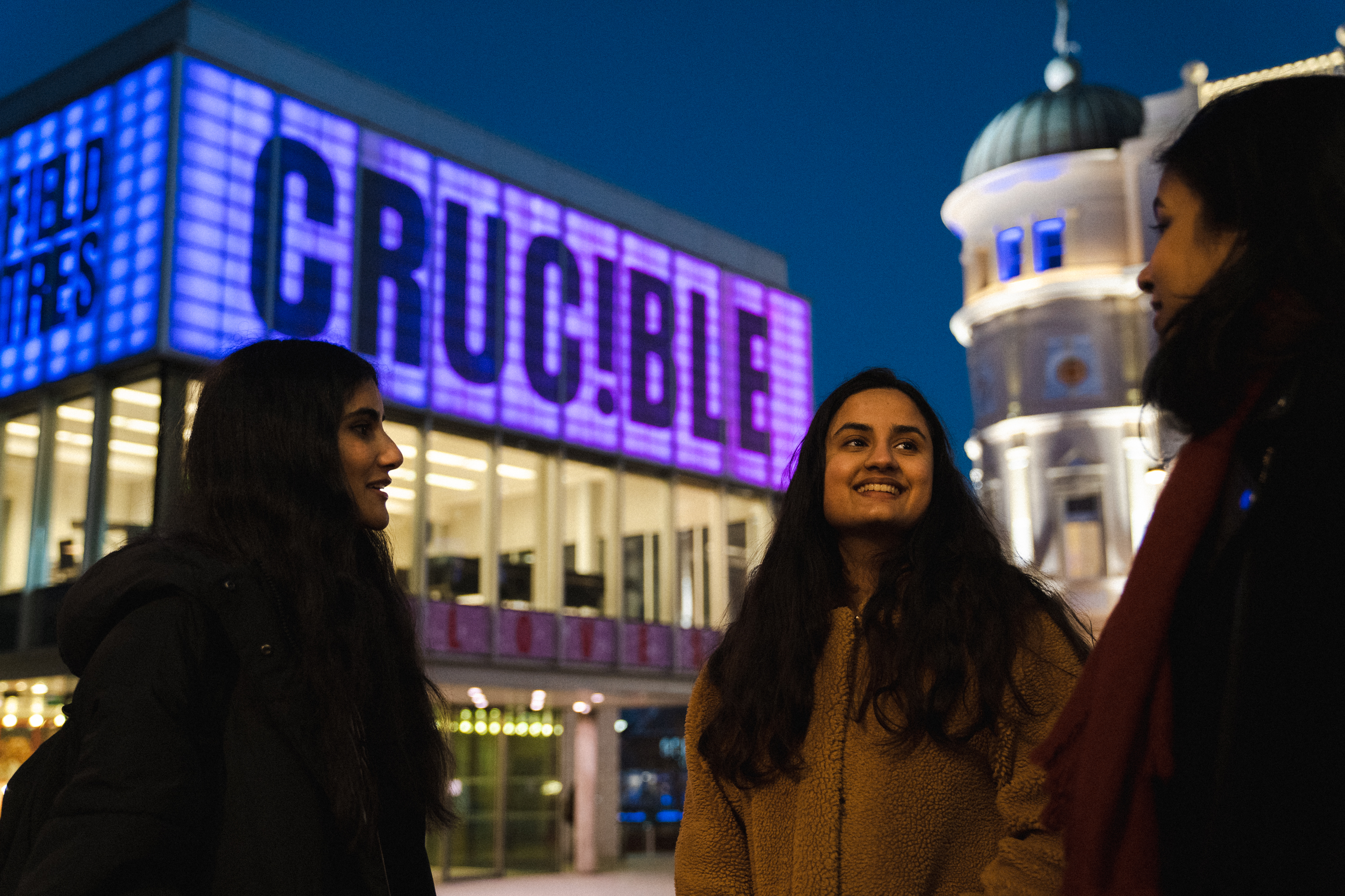 Khyati Mundrey Srivastav - University of Sheffield student: Three people standing outside the Crucible Theatre at night, with the building illuminated in purple and blue lights displaying the word “CRUCIBLE.” A historic domed building is visible in the background under a dark sky.
