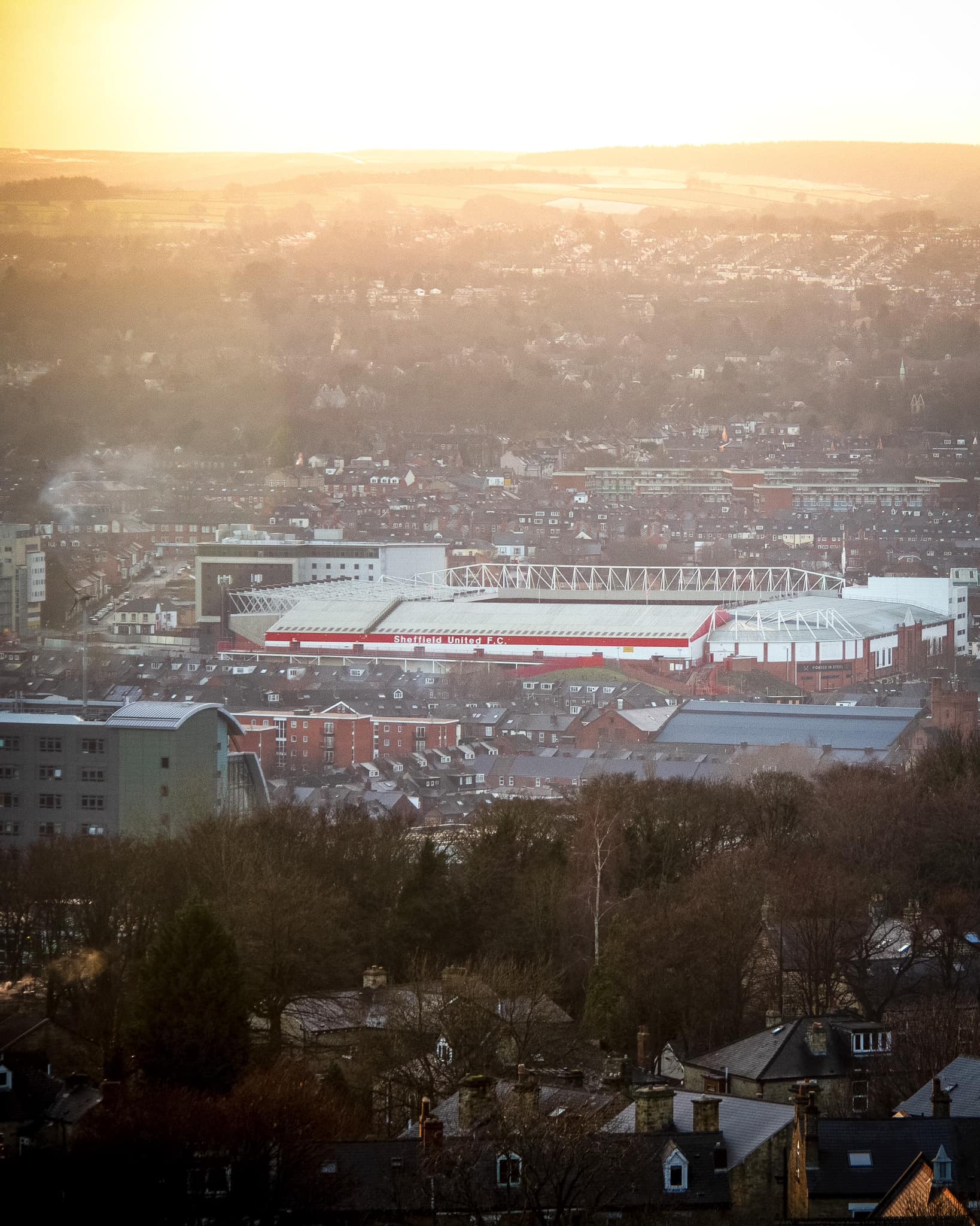 Aerial view of Bramall Lane stadium in Sheffield, surrounded by densely packed buildings and houses. The stadium has a white roof with red accents and is set against a backdrop of rolling hills under a bright, hazy sunset.