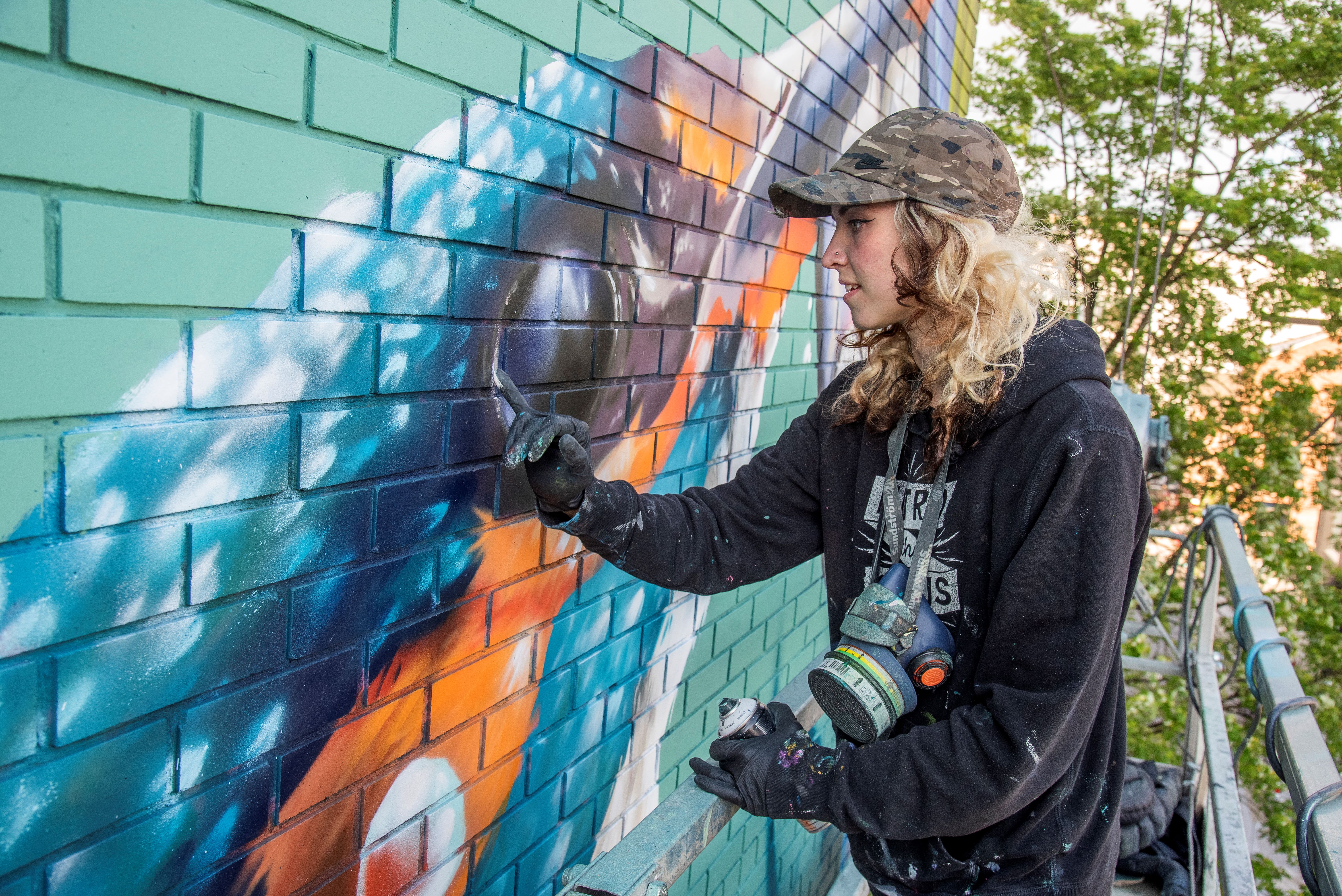 A Person wearing a dark hoodie and cap painting a colorful mural on a green brick wall using spray paint, with shades of blue, orange, and white forming abstract shapes. The scene includes a metal railing and leafy trees in the background.