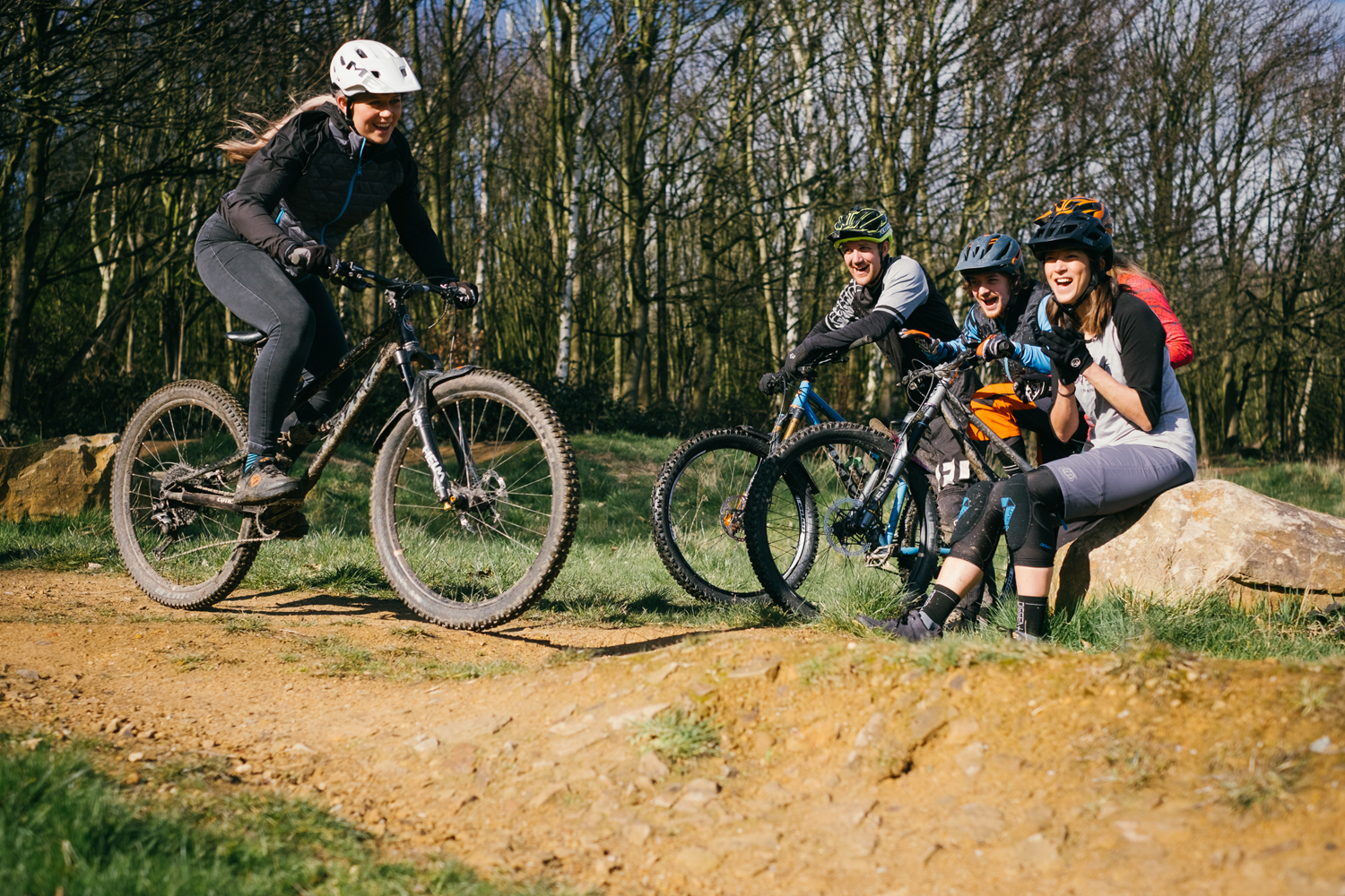 Female mountain biker riding on dirt track. Three other bikers are sat to the right on a log cheering her on.