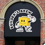 Builder's Brew Café logo on the outside of a building. The logo has the words 'Builder's Brew' round a cartoon character, who is shaped like a toy building brick, holding a mug of tea.