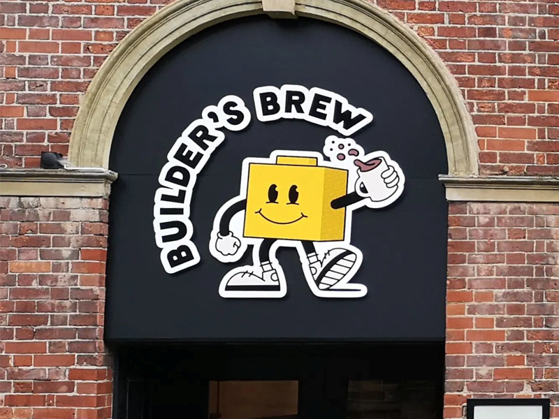 Builder's Brew Café logo on the outside of a building. The logo has the words 'Builder's Brew' round a cartoon character, who is shaped like a toy building brick, holding a mug of tea.