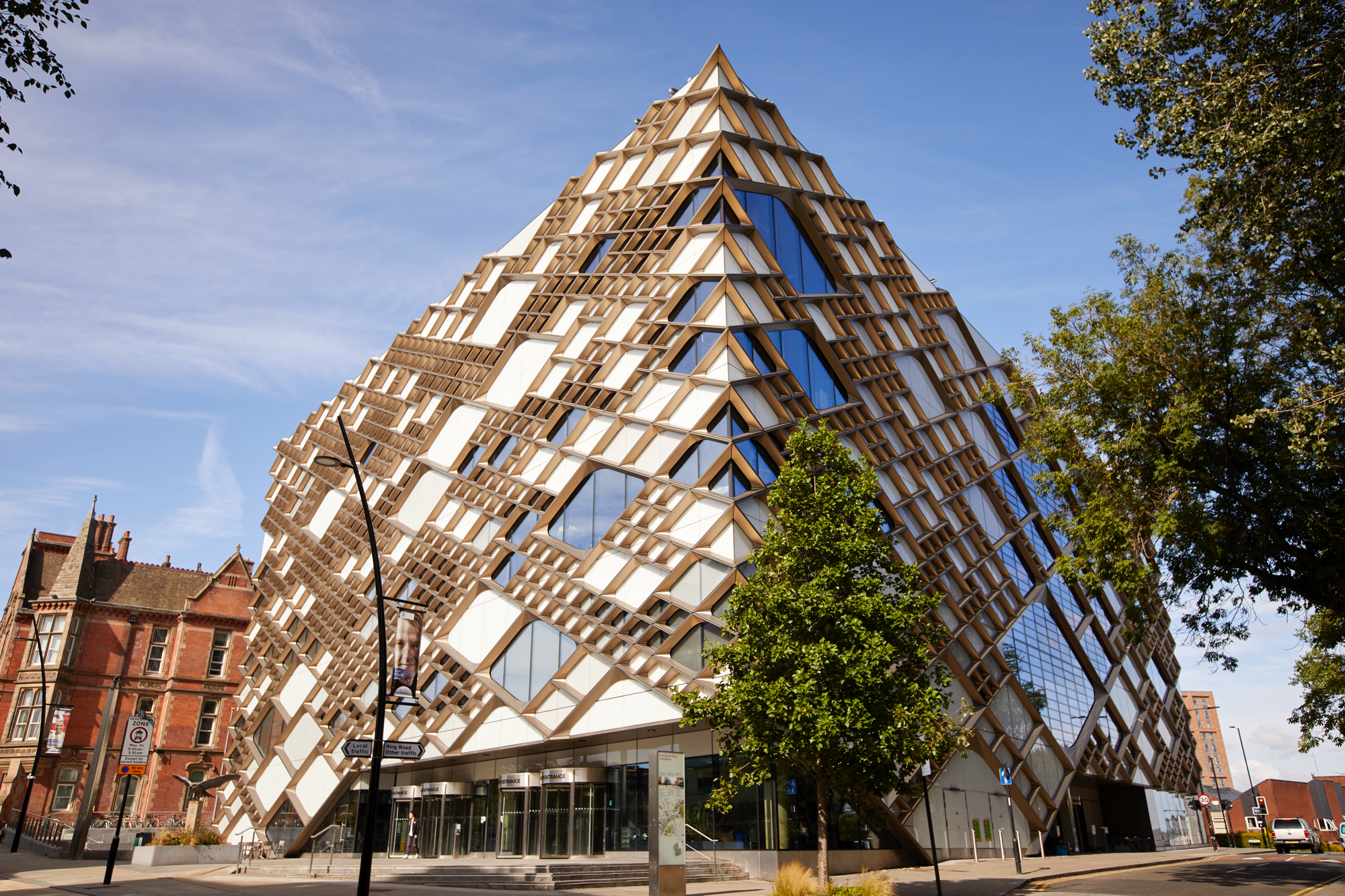 A modern, pyramid-shaped building with a striking geometric design featuring a mix of glass panels and wooden lattice structures. The facade reflects the blue sky, and the building is surrounded by trees and a few older brick buildings on the left. The entrance area has large glass doors, and the street in front includes signage and a lamppost.