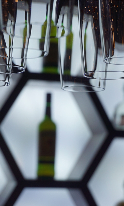 Close-up of an illuminated bar display featuring hexagonal shelves with bottles of liquor arranged inside. Several upside-down hanging glasses are in the foreground, creating a layered effect. The shelves have a bright white backlight, contrasting with the dark surroundings and highlighting the shapes of the bottles and glassware.