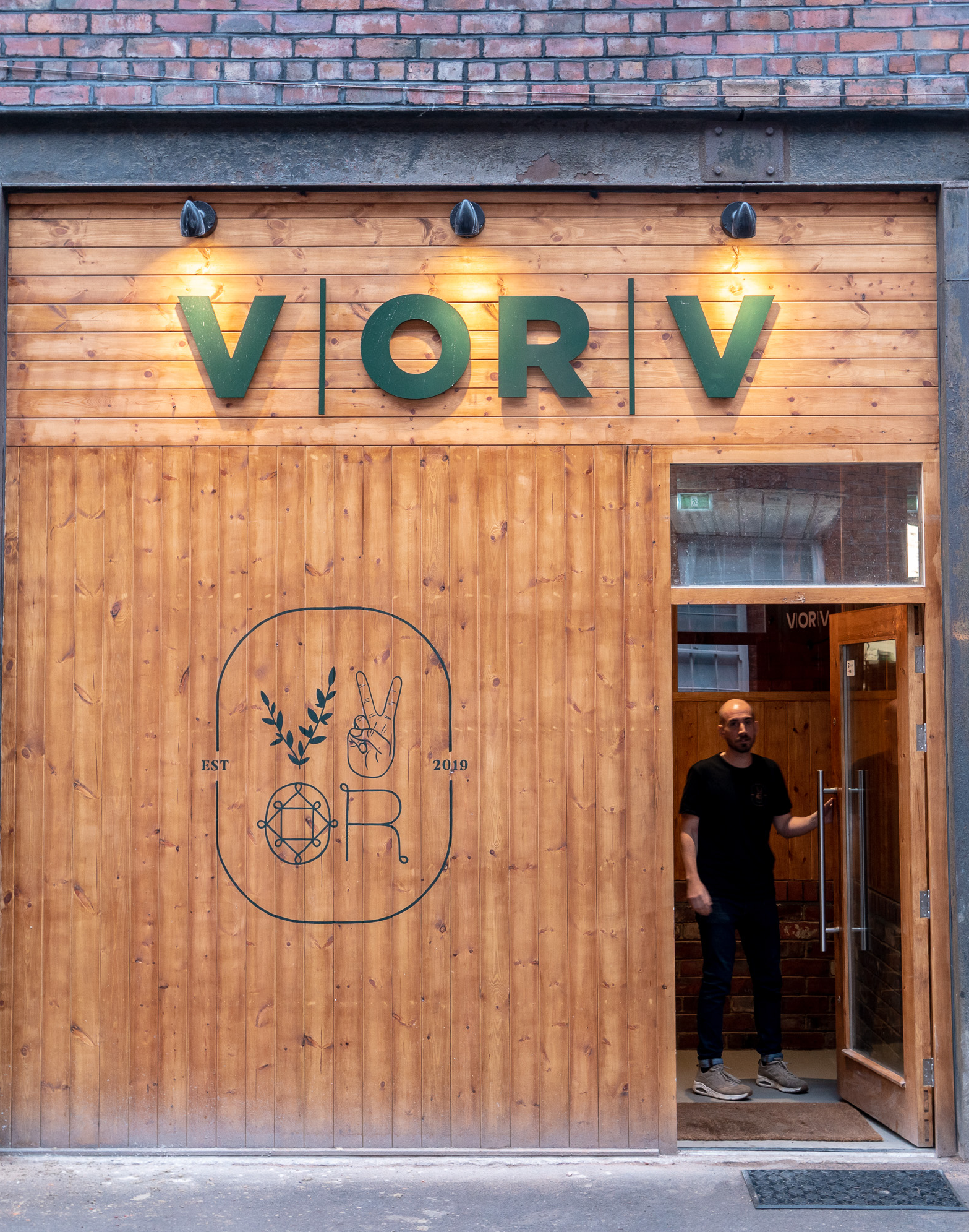 Exterior view of a wooden storefront with the name “V | OR | V” displayed in large green letters above the entrance. The facade features three wall-mounted lights and a logo with illustrations of leaves, a peace sign hand gesture, and geometric shapes, along with the text “EST 2019.” A person is standing in the doorway, which is open, revealing part of the interior. The building has a brick upper section above the wooden panelling.