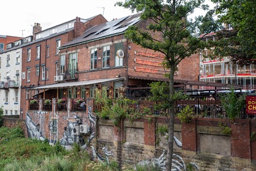 Brick building with a riverside terrace and outdoor seating, featuring a large neon sign on the upper wall. The lower wall is decorated with a monochrome mural of intertwined hands. Surrounding area includes trees, greenery, and adjacent buildings under an overcast sky.