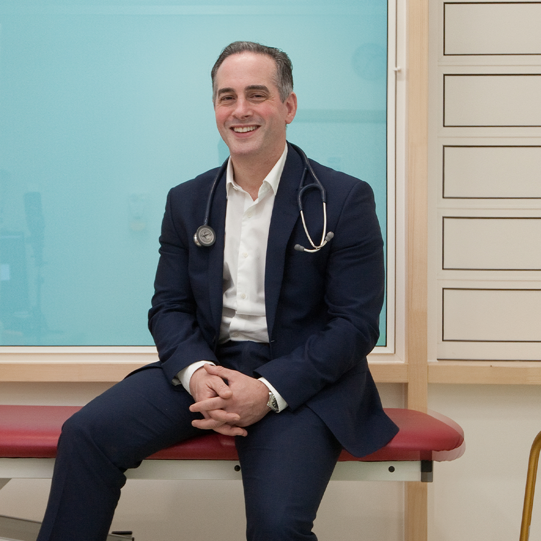 A person wearing a dark navy suit with a white dress shirt, seated on a red cushioned examination table in a clinical setting. A stethoscope hangs around the neck, suggesting a medical professional. The background includes a light blue frosted glass panel and a section of white drawers on the right side.