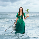 A woman, in a long green dress, is standing in the sea with waves rolling in towards her. In one hand she has a small stepladder, in the other a pot plant. Draped  around her shoulders is a power lead extension. She looks confused.