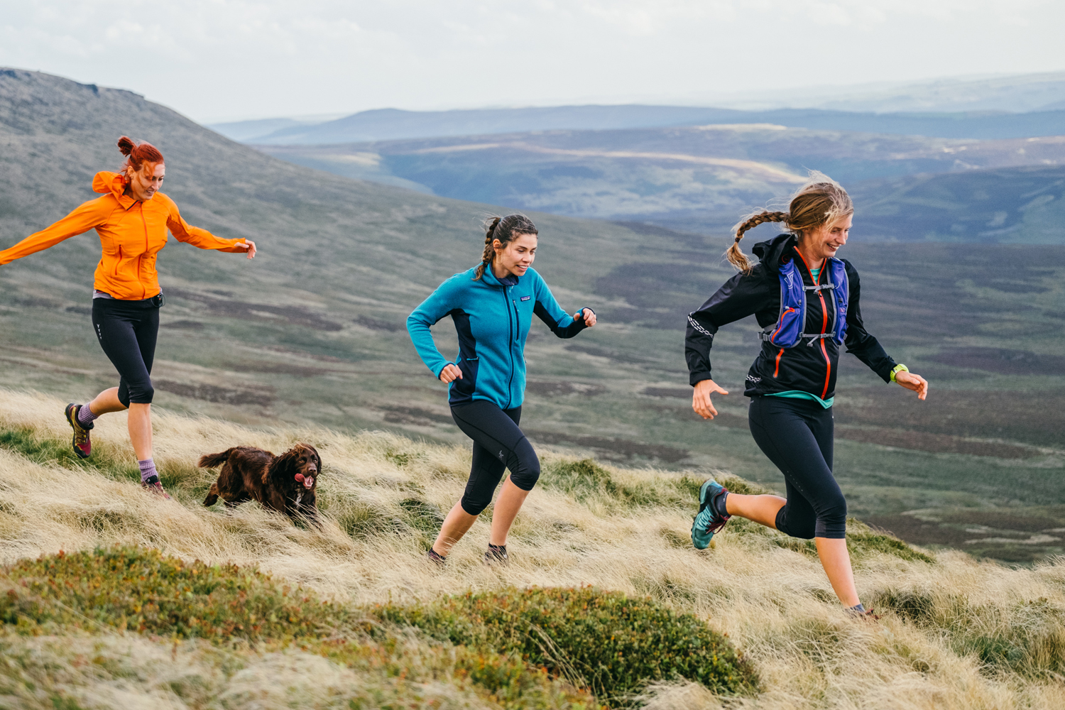 Three people running along a grassy hillside in an open moorland landscape, accompanied by a brown dog. The runners are wearing outdoor athletic clothing, including jackets and leggings, and the terrain is covered with tufts of grass and low vegetation. Rolling hills and expansive valleys stretch into the background under a partly cloudy sky.