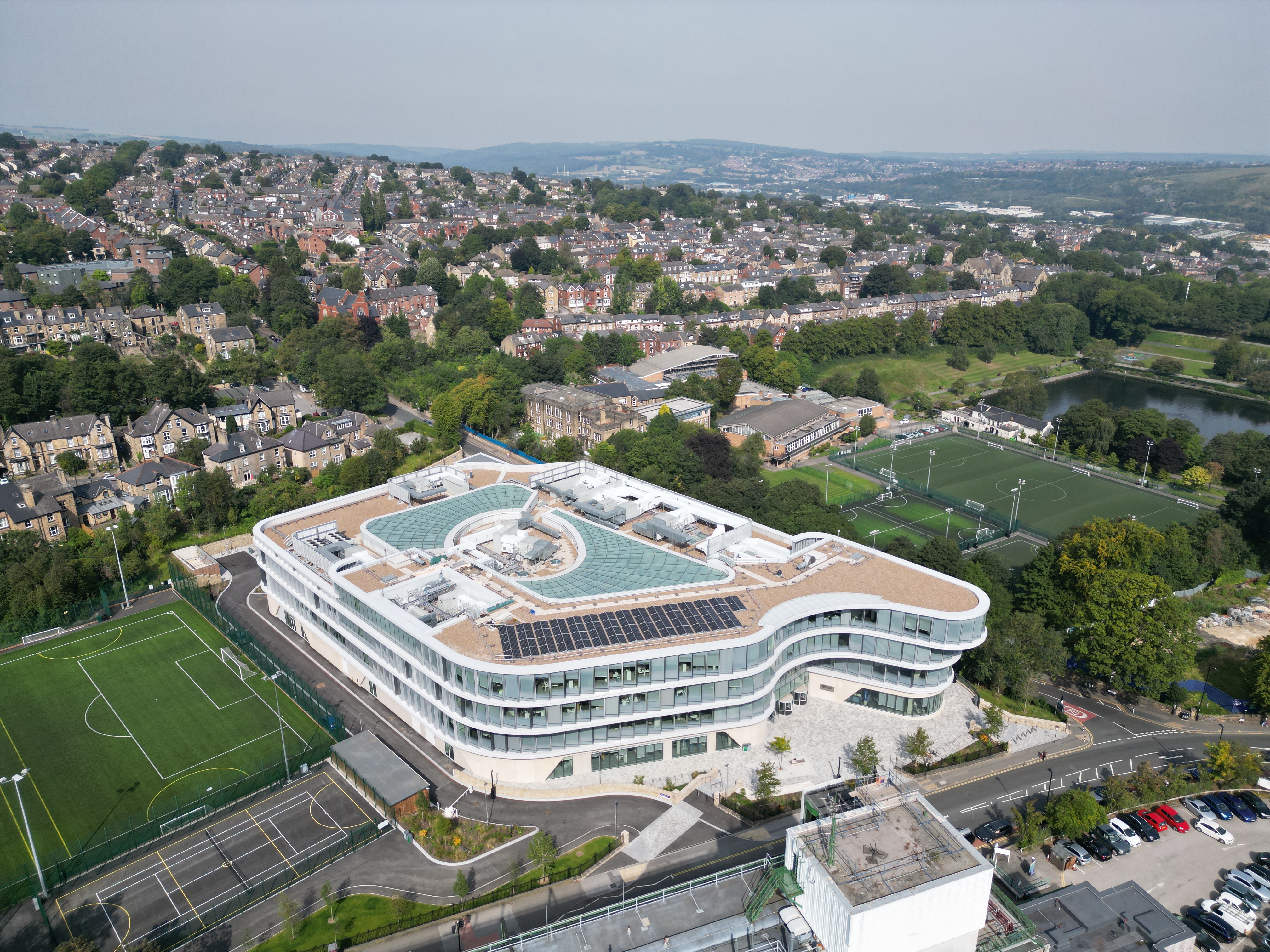 Aerial view of a modern, curved glass building with rooftop solar panels and a central circular feature, surrounded by sports fields and parking areas. In the background, a residential neighborhood with rows of houses and green spaces stretches across rolling hills under a hazy sky.