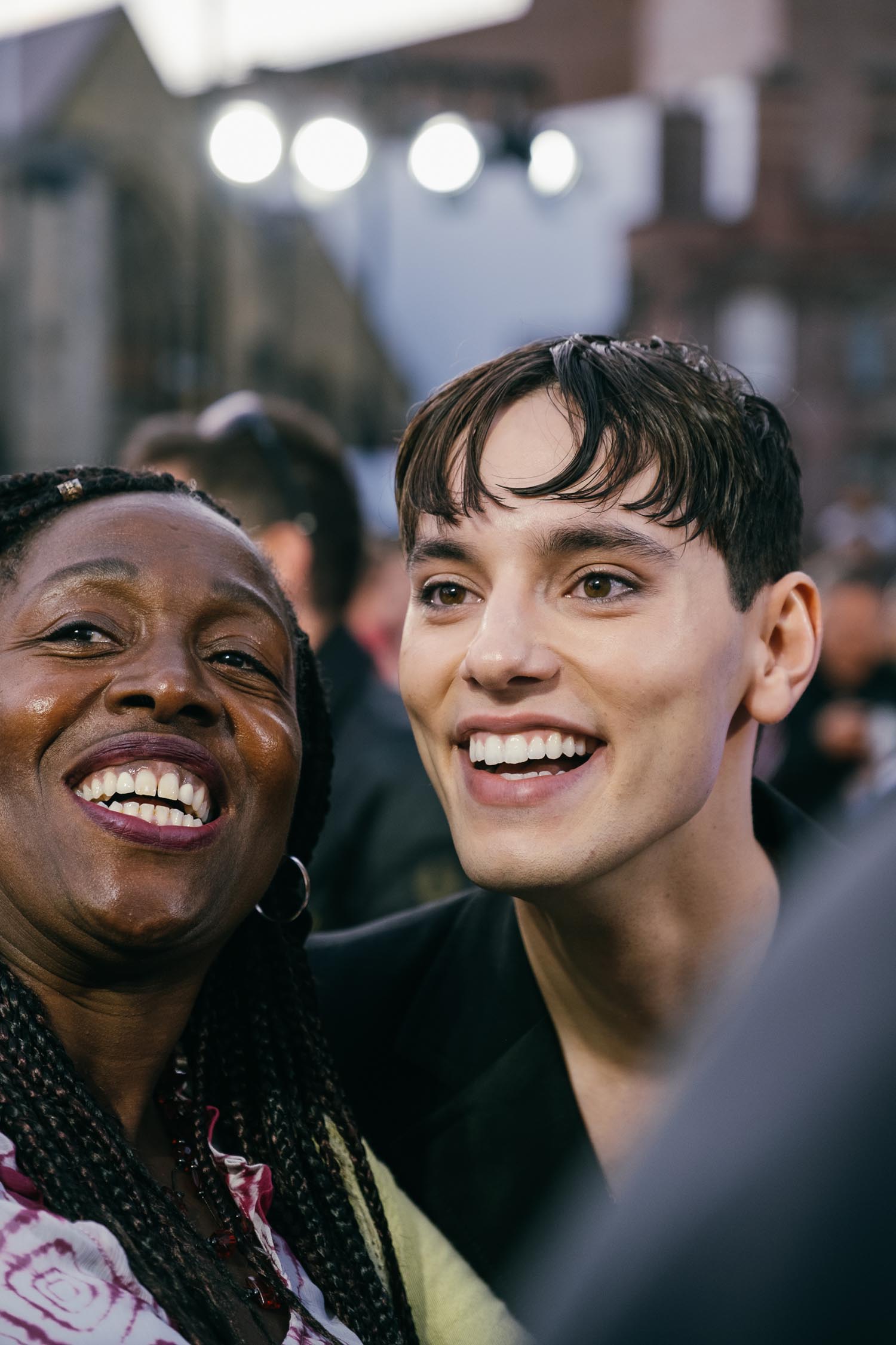 Two people standing close together in an outdoor setting, with a crowd and buildings visible in the background. The image shows bright stage lights overhead and blurred architectural details behind them, suggesting an event or gathering in an urban area. One person is wearing a dark outfit, while the other has braided hair and a patterned top.