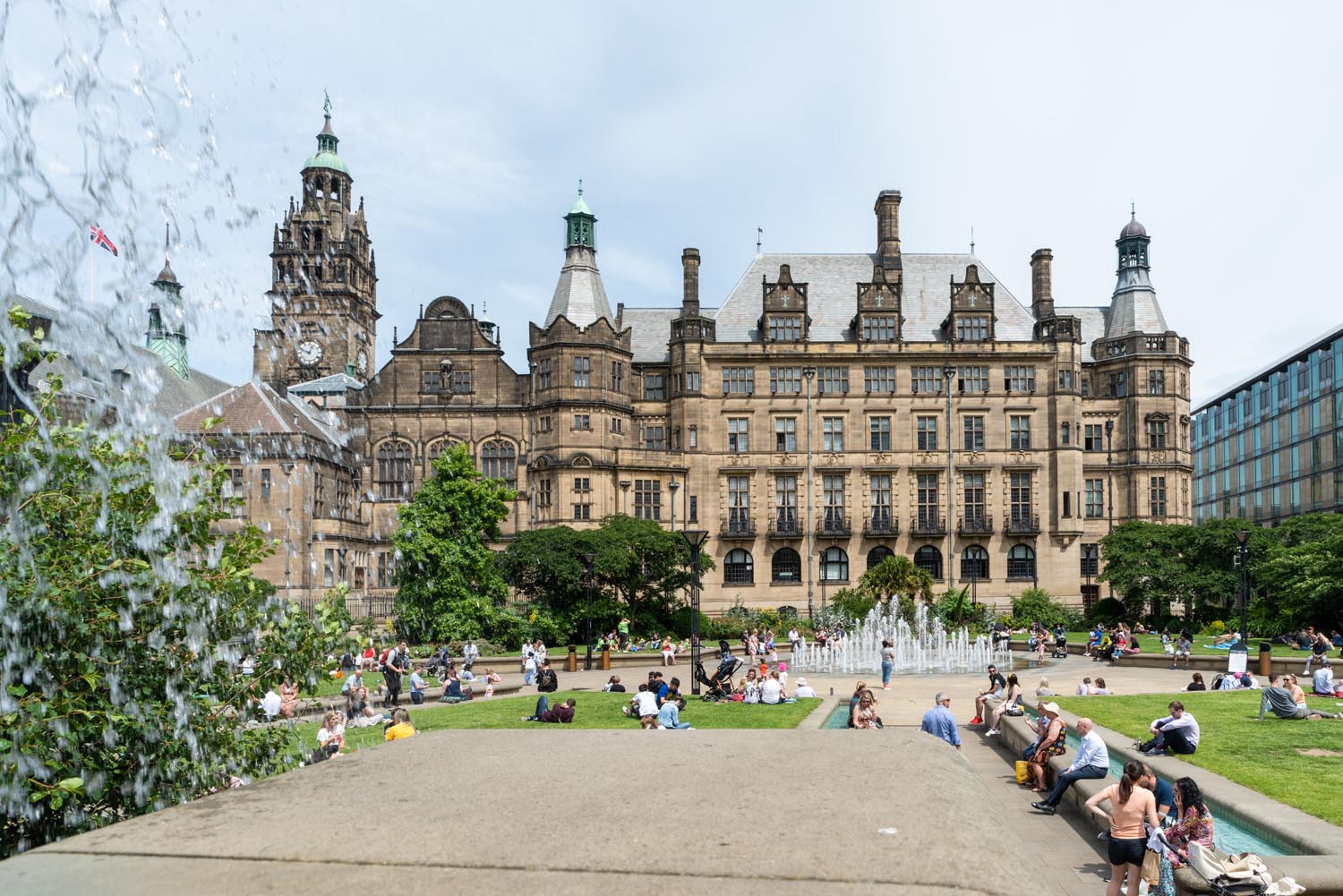 View of Sheffield Town Hall from the Peace Gardens, showing the historic stone building with ornate towers and spires. In the foreground, people are sitting and relaxing around fountains and green lawns under a bright sky.