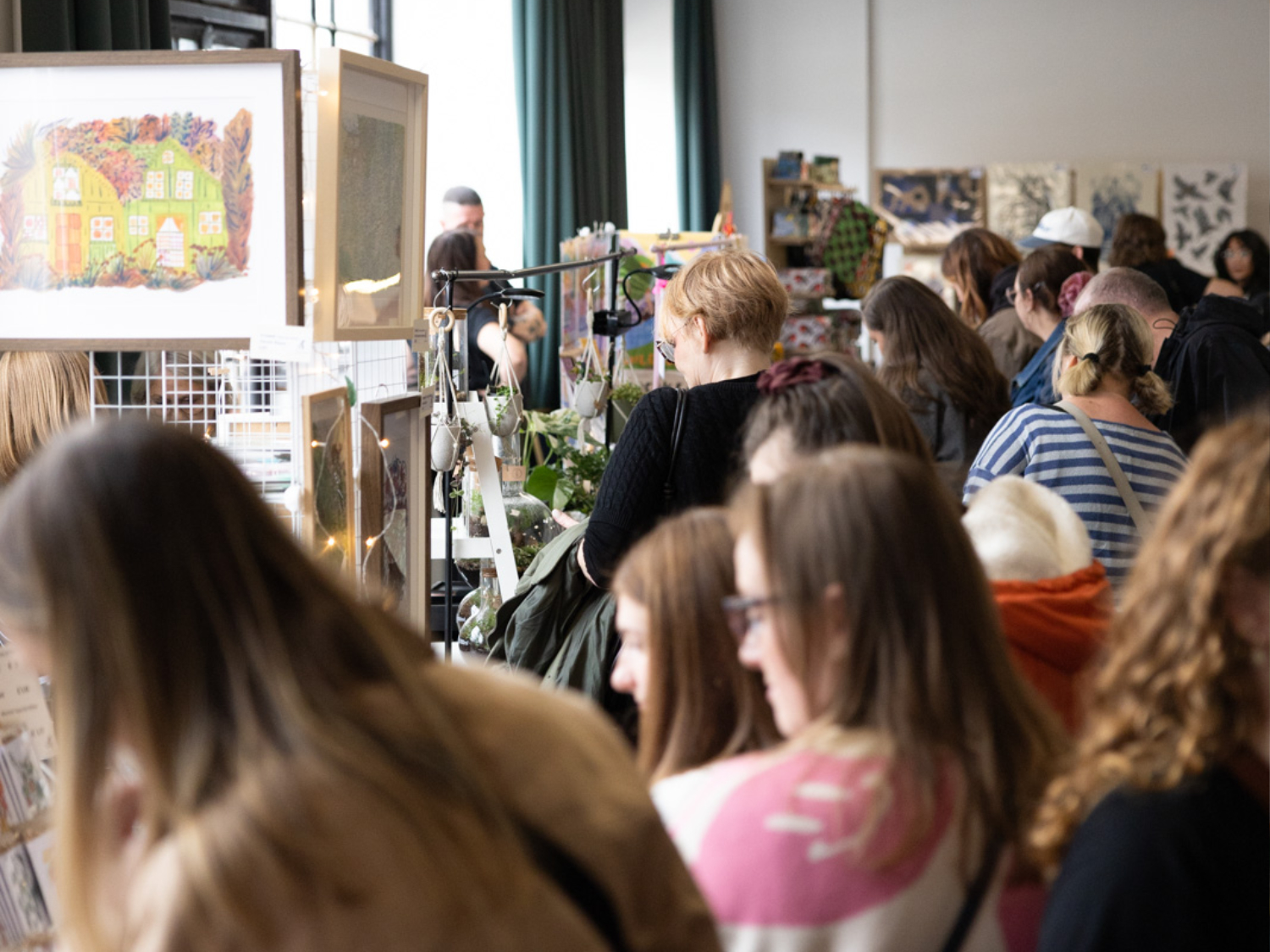 A busy indoor market or fair with people browsing stalls selling artwork, crafts, and plants. A framed painting of a house with colorful foliage is prominently displayed. The scene captures a lively community event focused on local art and handmade goods.