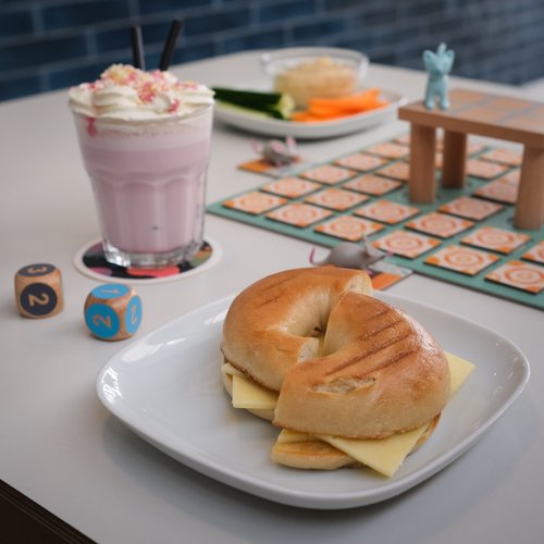 A bagel on a plate and a milkshake, sat on a table that also has a board game set up on it.