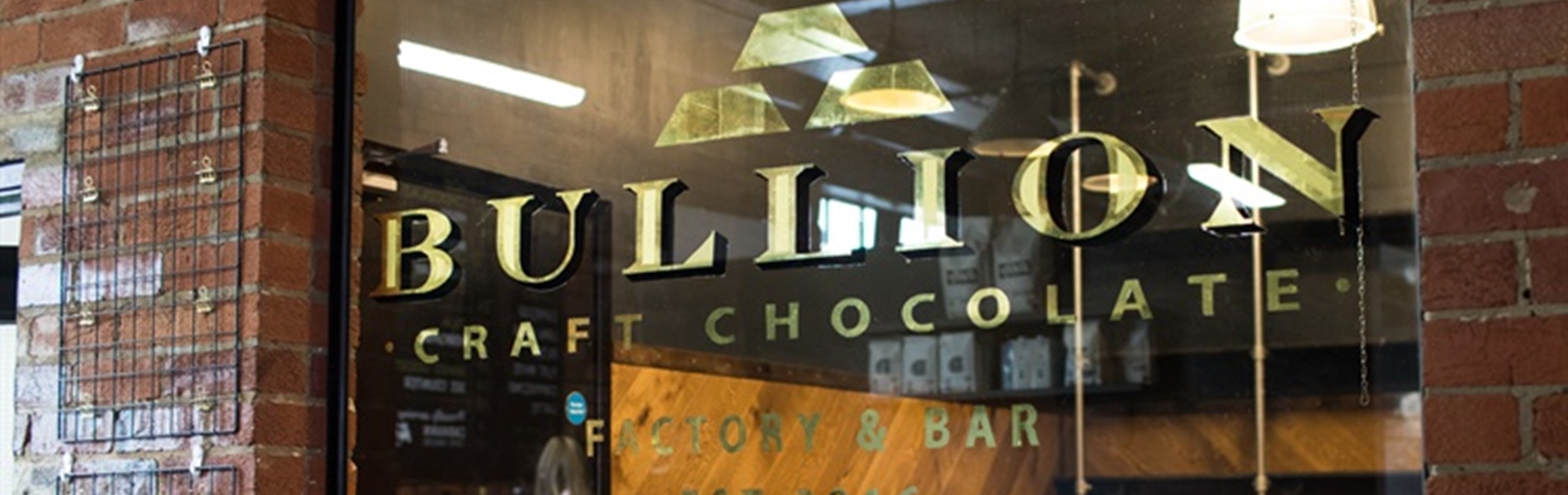Storefront of Bullion Craft Chocolate, featuring a glass window with the gold Bullion logo and name. Inside, wooden shelves, a ladder, and hanging lights are visible. A person is seen through the window. The building has a red brick exterior.