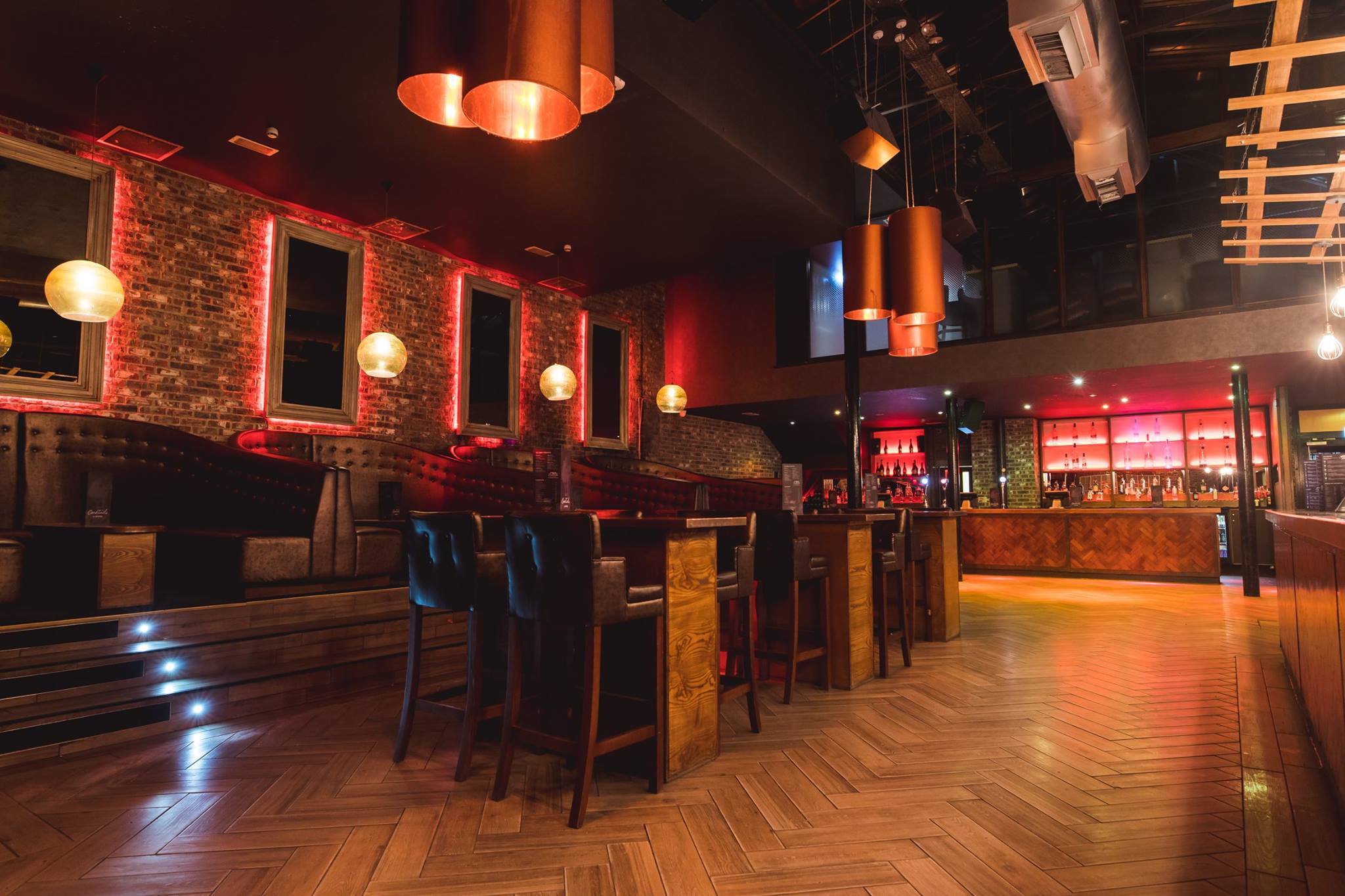 Interior shot of Crystal. Modern bar interior with warm lighting, exposed brick walls, and wooden herringbone flooring. The space features high tables with leather chairs, booth seating along the wall, and a long bar counter with illuminated shelves stocked with bottles. Copper pendant lights hang from the ceiling, adding to the stylish atmosphere.