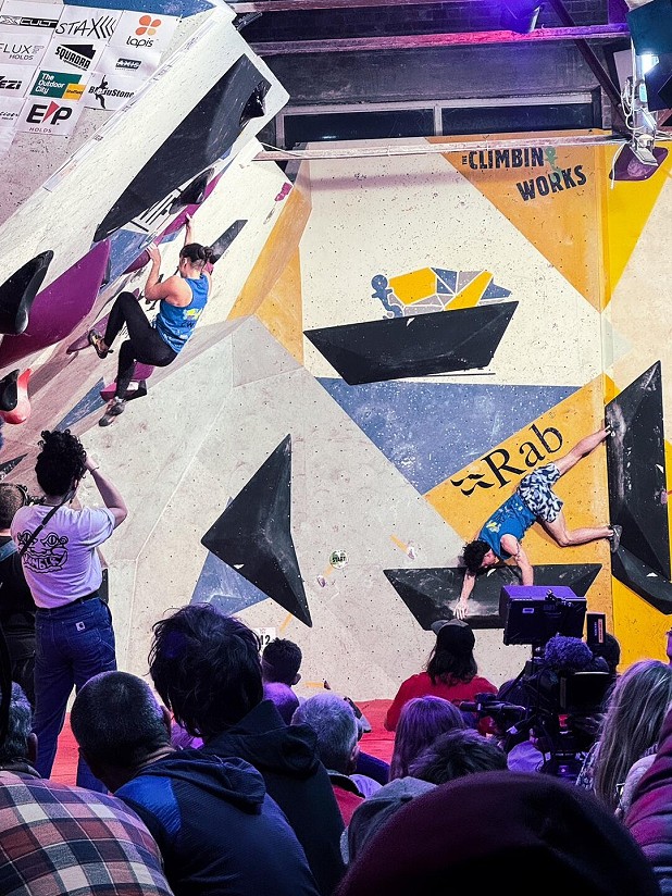 Two climbers wearing blue tops compete on an indoor bouldering wall with large black and purple holds. The wall features bold geometric panels in yellow, white, and blue with visible event branding, including “Rab” and “Climbin Works.” A crowd of spectators and camera operators watch from the foreground under bright stage lighting.