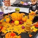 Festive Halloween-themed setup featuring a carved pumpkin centerpiece surrounded by small pumpkins, gourds, and autumn leaves. Children play around the table, including one in a skeleton costume placing items into a yellow bucket with a smiling face. The scene is vibrant and captures the spirit of fall celebrations.