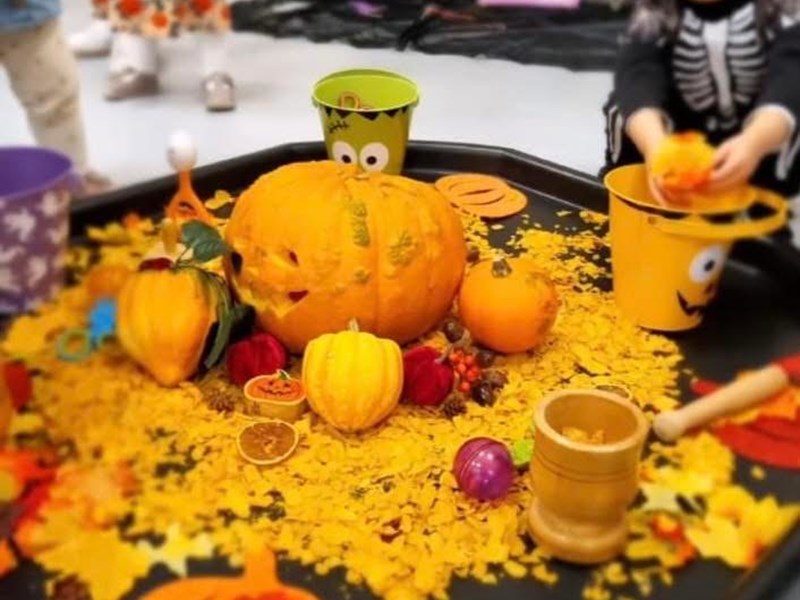 Festive Halloween-themed setup featuring a carved pumpkin centerpiece surrounded by small pumpkins, gourds, and autumn leaves. Children play around the table, including one in a skeleton costume placing items into a yellow bucket with a smiling face. The scene is vibrant and captures the spirit of fall celebrations.