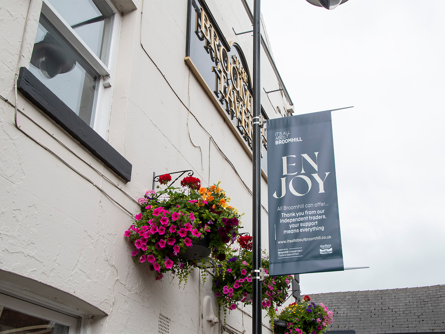 Exterior wall of a light-colored building with hanging baskets filled with vibrant flowers in pink, purple, and red. A vertical banner attached to a pole reads “ENJOY” and promotes local independent traders in Broomhill, with a website link at the bottom. Part of the building sign for “Broomhill Tavern” is visible above the baskets, and rooftops appear in the background under a cloudy sky.