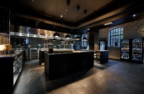 Modern professional kitchen with dark flooring and black cabinetry. The space features large stainless steel counters, multiple black pendant lights, and industrial ventilation ducts overhead. A wall with large windows and exposed brick is visible on the right, alongside refrigerators stocked with bottled drinks. Cooking equipment and utensils are neatly arranged throughout the kitchen.