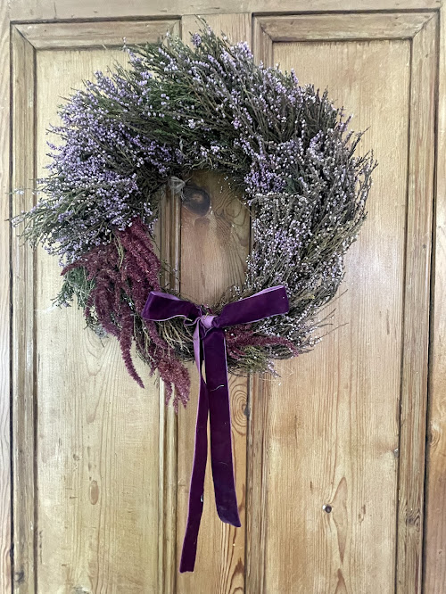 A rustic wreath made of dried purple heather and deep burgundy foliage, accented with a rich purple velvet ribbon tied in a bow. The wreath hangs on a natural wooden door, creating a warm and earthy aesthetic.