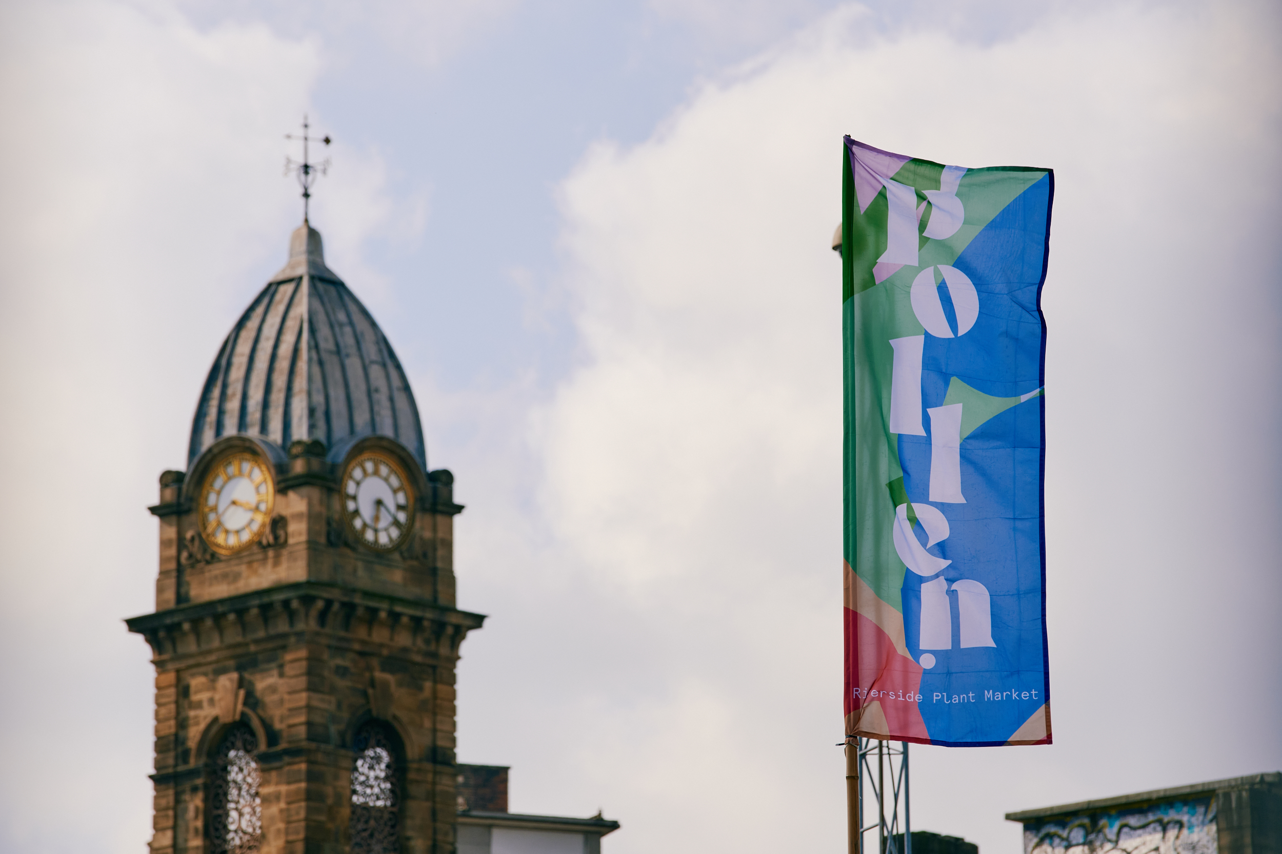 In the foreground is a colourful flag that has the word 'Pollen' on it, in the background is a clock tower.