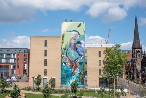 Large mural painted on the side of a tall brick building depicting vibrant wildlife, including a heron, a kingfisher, butterflies, and reeds in a water scene. The building is surrounded by green trees and a landscaped area, with modern apartments on the left and a historic church with a tall spire on the right under a partly cloudy blue sky.