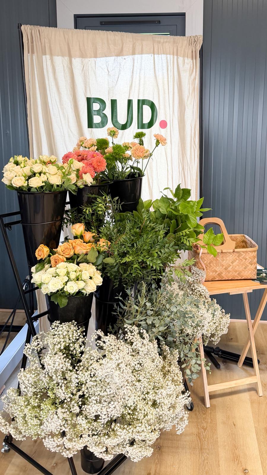 Indoor floral display with multiple black vases holding assorted flowers and greenery, including white baby’s breath, cream and peach roses, and pink carnations. A wicker basket sits on a small wooden stand to the right. Behind the arrangement is a beige curtain with the word “BUD.” printed in green and pink letters. The setting features a wooden floor and dark panelled walls.
