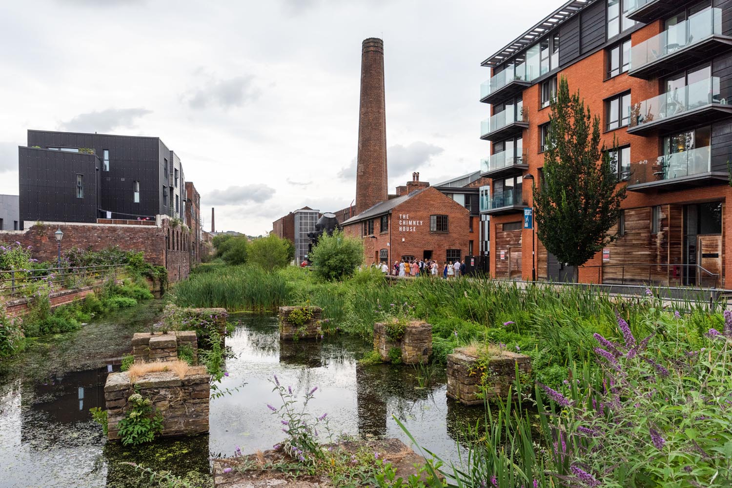 Building, a mixture of old and new, stand along the banks of a river. In the distance is a chimney stack rising up out of Kelham Island Museum.